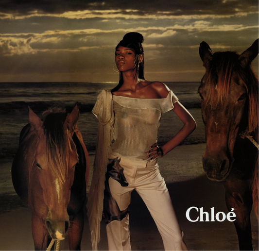 Chloé campaign of model in ivory one-shoulder pinstripe top between horses on a beach